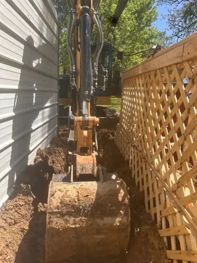 A mini excavator bucket opening a narrow trench along the side of a house, between the siding and a wooden lattice fence