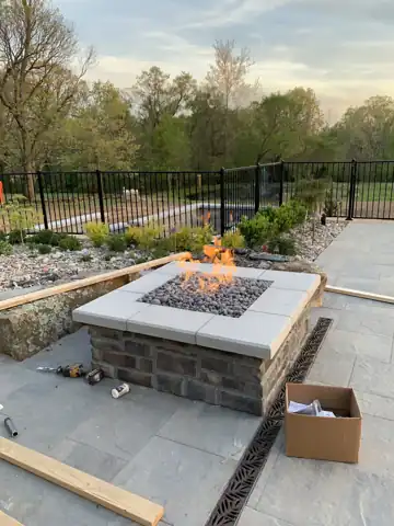 An outdoor gas fire pit on a stone patio, lit, with construction materials still around it.