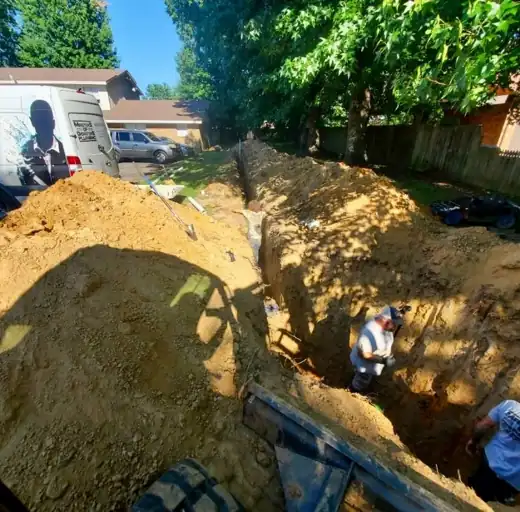 A crew member working at the bottom of a deep water line trench in a back yard, the dirt piled high beside it
