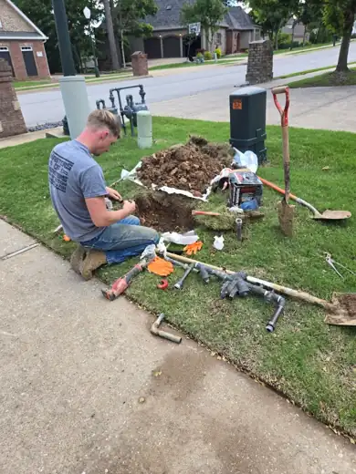 A plumber kneeling at an opened water meter box on a front lawn, new fittings and tools laid out on the grass