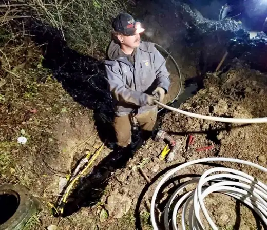 A technician with a headlamp pulling white PEX through a trench at night.