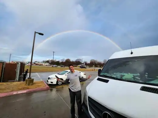 A Masters of Disasters crew member smiling beside the company van under a double rainbow in a parking lot